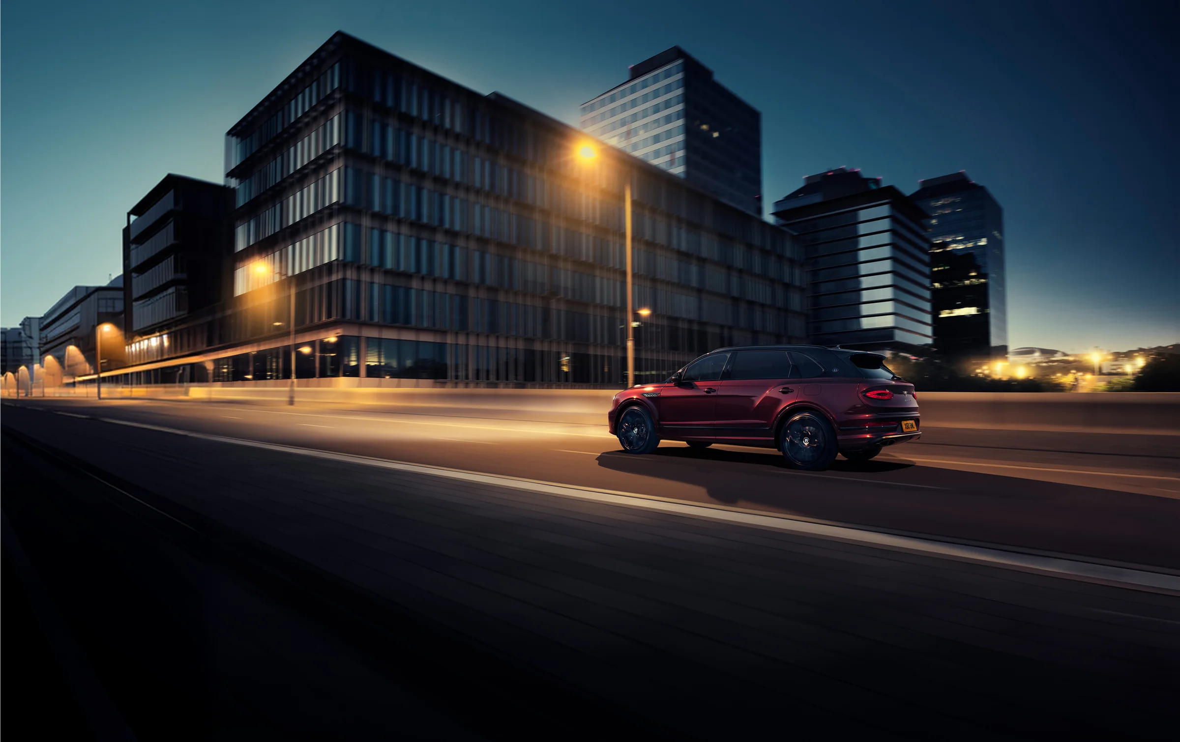 A Bentley Bentayga in dark red on a city road at dusk, the buildings behind it lit and blurred.