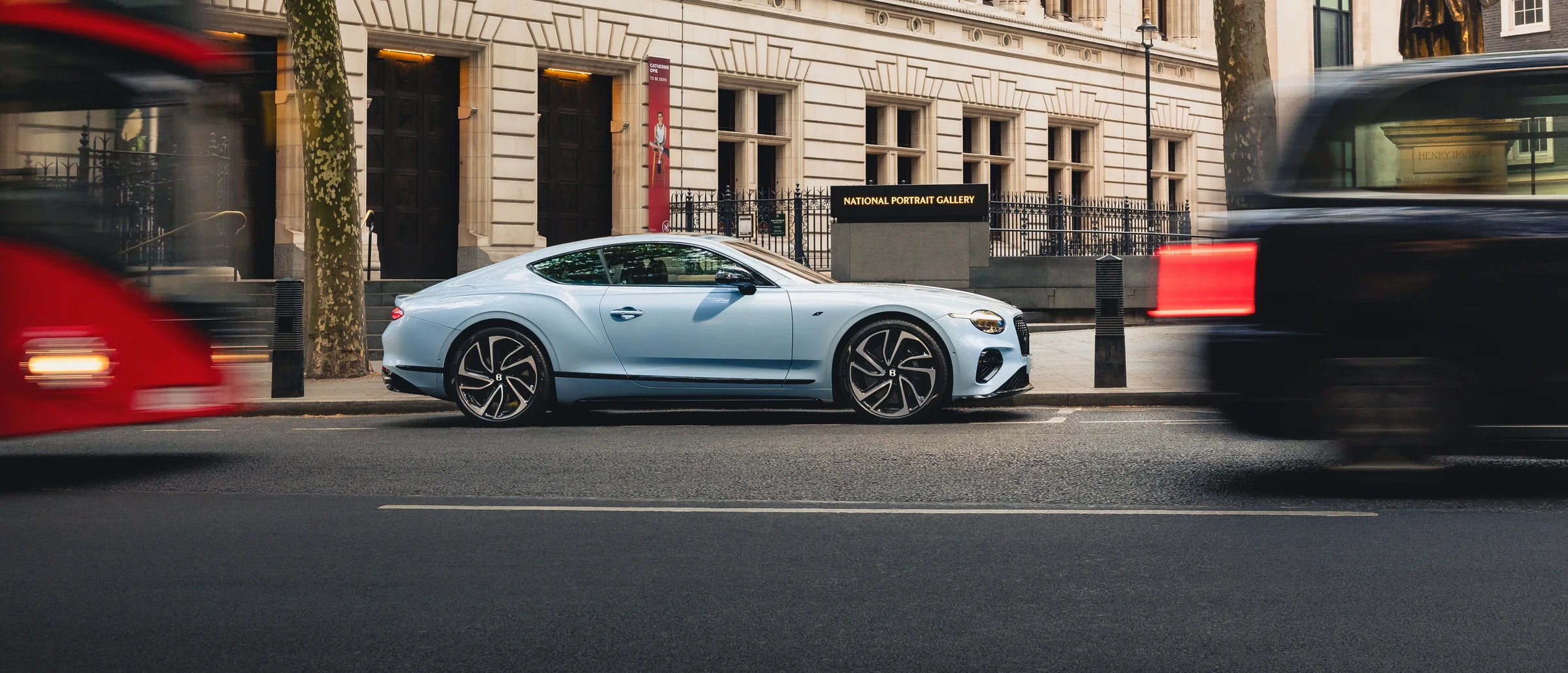 A Bentley Continental GT in pale blue at rest on a city street outside a stone gallery building, traffic passing in front of it.